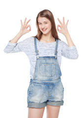 Happy young fashion girl in jeans overalls gesturing okay isolat