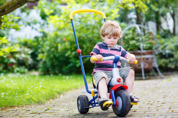 Active blond kid boy driving tricycle or bicycle in domestic gar
