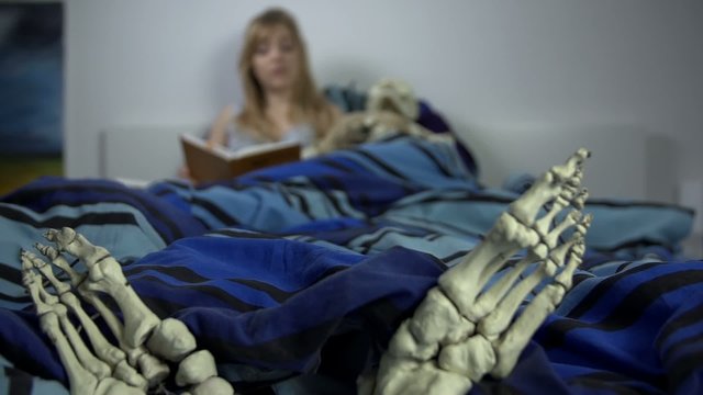 Woman reads in the bed with the skeleton laying beside her