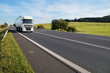 Asphalt road in a rural landscape, the arriving two white trucks