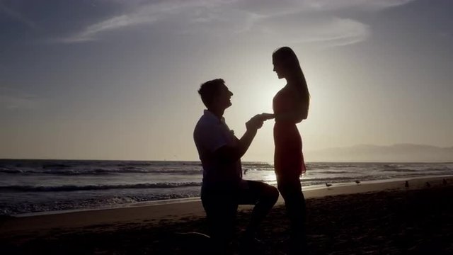 man proposing marriage to woman on one knee - will you marry me - silhouette on beach by ocean - romantic proposal putting ring on finger to be married - 4K slow motion