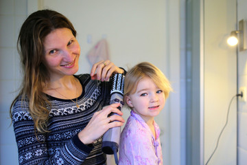 Young mother helps daughter to style hair with round brush dryer
