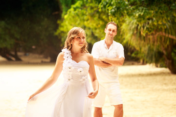 bride and groom stand on sand beach barefoot near green trees