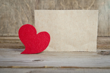 Red fabric heart with sheet of paper standing on a wooden board.