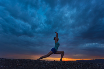 Woman practicing Warrior yoga pose outdoors over sunset sky back