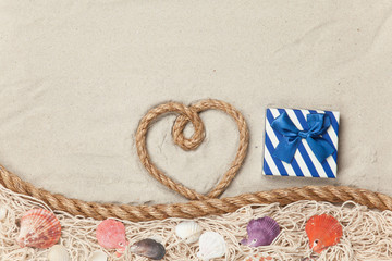 Fototapeta na wymiar Gift box and rope in heart shape near net and shells