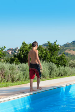 Boy standing at the poolside. Children 10 years old walk around the edge of the pool in swimsuits. He can only be seen from behind. It is summer.