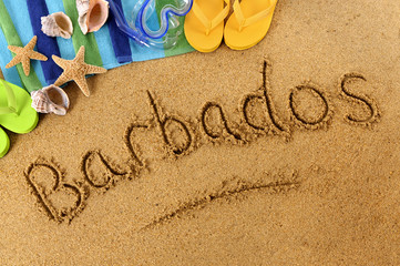 The word Barbados written in sand on a beach with towel flip flops seashells summer vacation holiday photo - obrazy, fototapety, plakaty