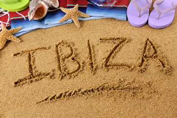 The word Ibiza written in sand on a beach with towel flip flops seashells summer vacation holiday...