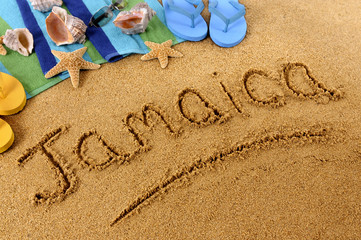 The word Jamaica written in sand on a beach with towel flip flops seashells Caribbean summer vacation holiday photo - obrazy, fototapety, plakaty
