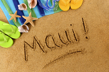 Fototapeta na wymiar The word Maui written in sand on a beach with towel flip flops seashells Hawaii summer vacation holiday photo