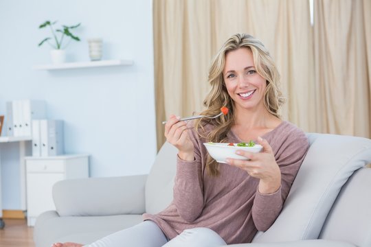 Smiling blonde sitting on couch eating salad