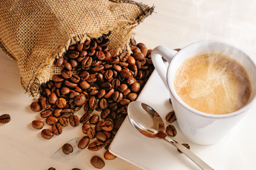 Cup of hot coffee on table and sack with coffee beans closeup