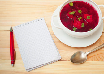 Red soup with dill in white bowl on wooden table with notebook f