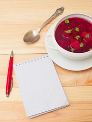 Red soup with dill in white bowl on wooden table with notebook f