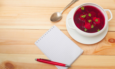 Red soup with dill in white bowl on wooden table with notebook f