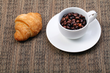 Coffee cup with a croissant and fresh coffee beans on a brown ba