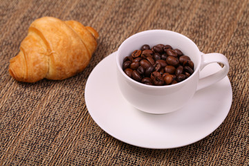 Coffee cup with a croissant and fresh coffee beans on a brown ba