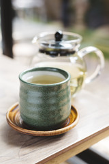Green tea cup on wooden table with glass tea pot in background