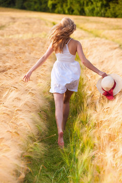 Happy young blonde girl in white dress with straw hat running th