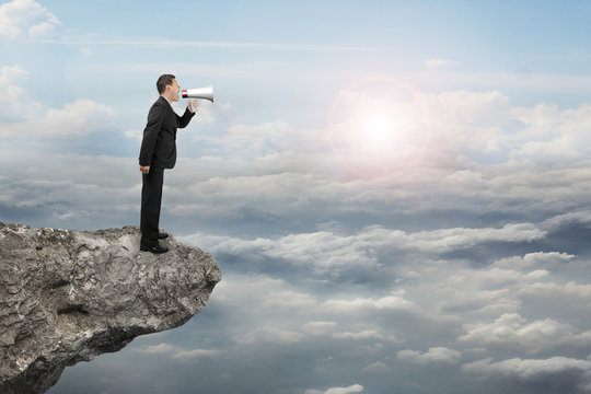 Businessman using megaphone yelling on cliff with sunlight cloud