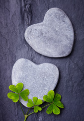 Fototapeta premium Two grey heart shaped rocks with three lucky clovers on tile ba