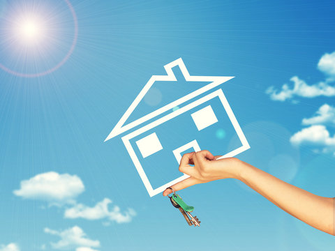 Hand holding house icon and key. Background of sky, clouds, sun