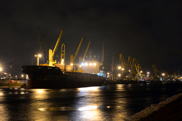 Fototapeta na wymiar The loading cargo ship with cranes is moored in port at night
