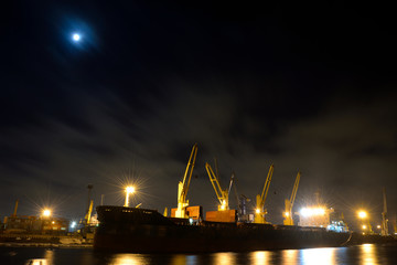 Fototapeta na wymiar The loading cargo ship with cranes is moored in port at night