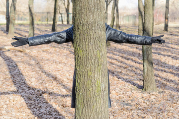 A woman wearing a black leather coat and gloves is hidden behind an oak tree in the park and spread her arms like a cross.
