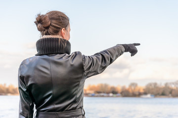 Fototapeta premium An adult with a chignon and wearing a black leather coat stays close to the river and point her finger to indicate something interesting on the opposite bank