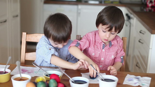 Two adorable boys, coloring eggs for Easter at home, having fun