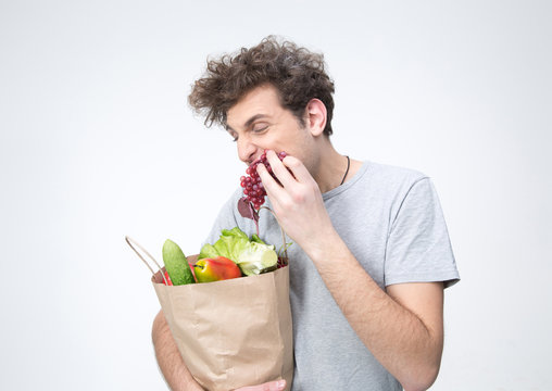 Handsome Man Holding A Bag Full Of Groceries And Eating Grapes