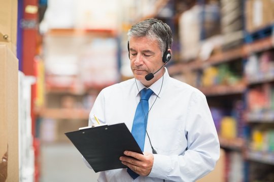 Warehouse manager wearing headset writing on clipboard