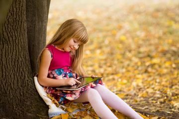 Little girl sitting on grass and leaf under tree and playing tab