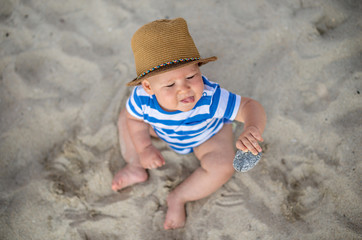 Baby boy with hat sitting on the beach.