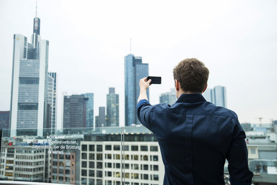 Germany, Hesse, Frankfurt, young man taking a picture of the skyline with his smartphone
