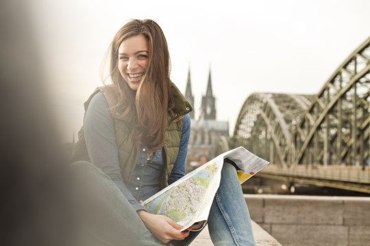Germany, Cologne, portrait of smiling young woman with city map on knee