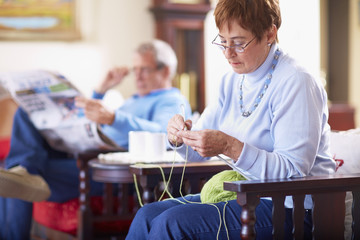 Senior woman knitting with husband in background reading newspaper