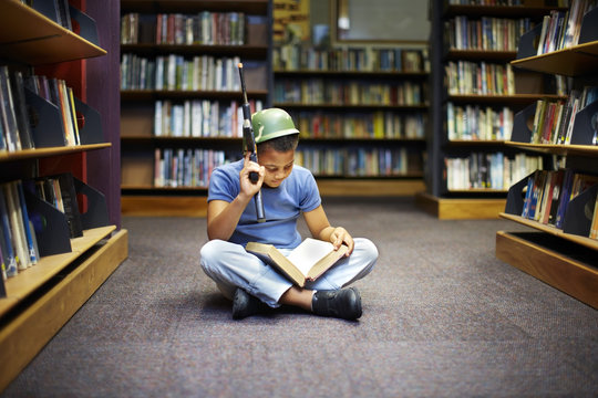 Boy with helmet and gun reading book in library