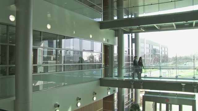 Backlit businessman and woman cross a walkway in a modern building atrium against a backdrop of white windows. Wide shot from same level as people.