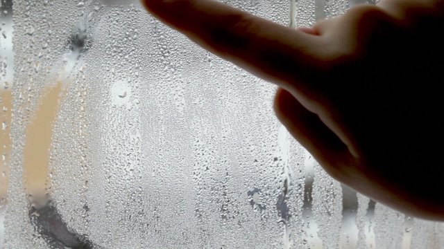 Childish hand draws smiling face by finger on wet glass, closeup