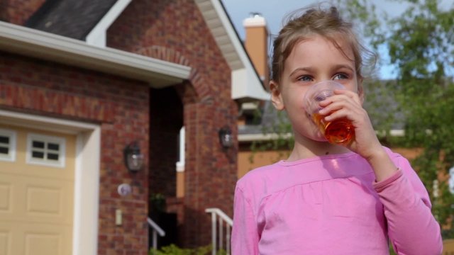 Little girl drinks juce and smiles near cottage at summer day