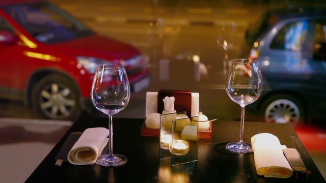 Candle burns at table with glasses and chopsticks, cars parked near road behind window
