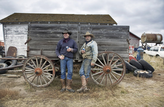Two men in cowboy hats and cowboy boots leaning against a wooden wagon.