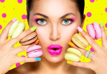 Girl with colorful makeup and manicure taking colorful macaroons - obrazy, fototapety, plakaty