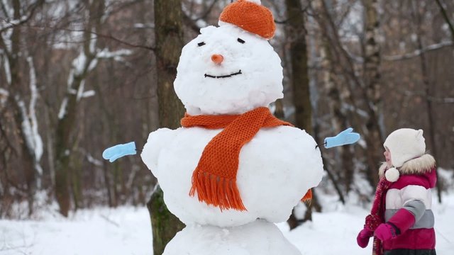 Little girl walk around snowman in orange hat and scarf 