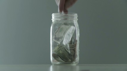 Hands put money into a tip jar or home savings, filling up the jar with dollars.