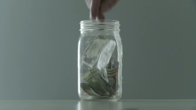Hands put money into a tip jar or home savings, filling up the jar with dollars.