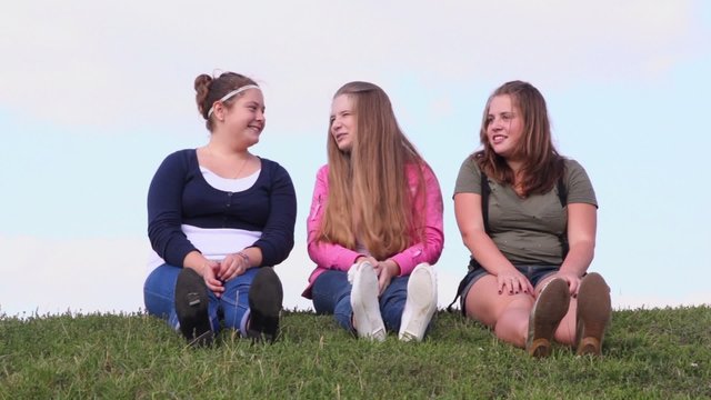 Three girls sit and talk on grass hill at sunny summer day